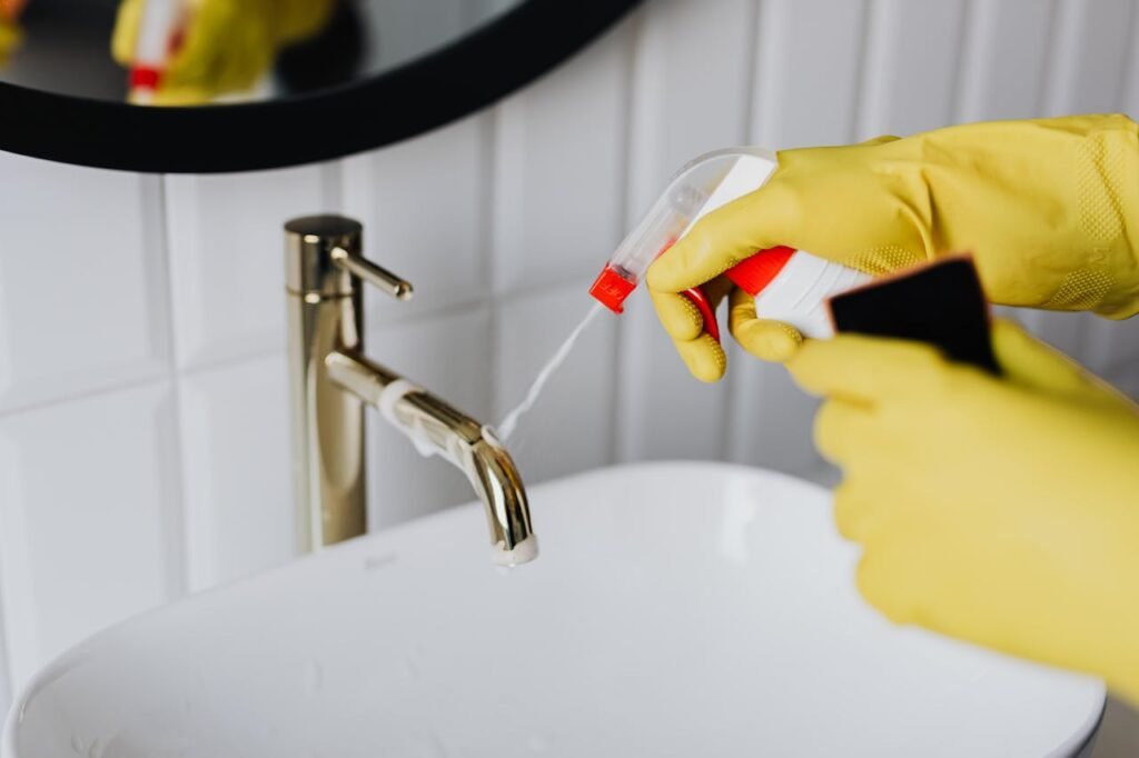 From above anonymous housekeeper in yellow rubber gloves spraying cleaner to chrome water faucet in bathroom, detergent factory